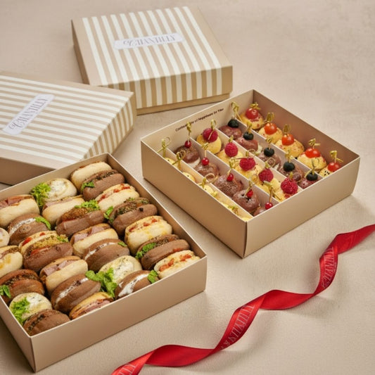 Two open boxes of assorted mini sandwiches and pastries with a red ribbon on a beige background.