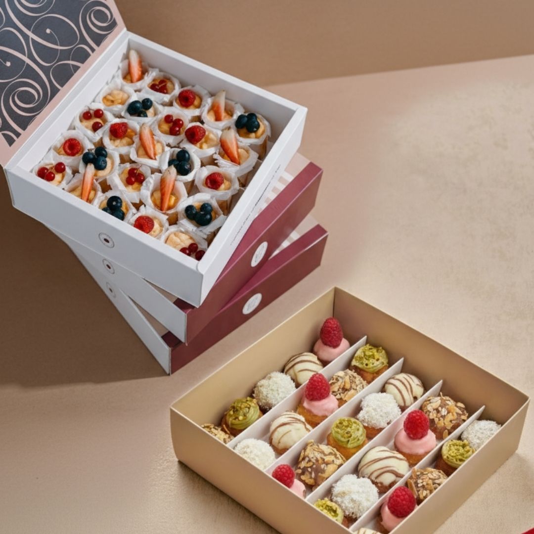 Two boxes of assorted small desserts on a beige surface